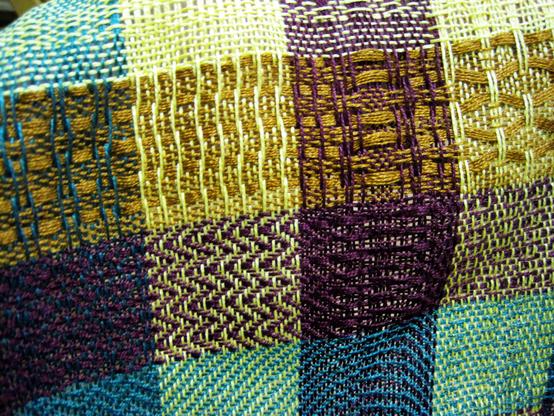 A close-up photograph of a threaded woolen fabric with squares of different colors and threads of color that overlap across the squares. By Candice under a CC BY-NC-ND license: https://bit.ly/3TW0hG7.