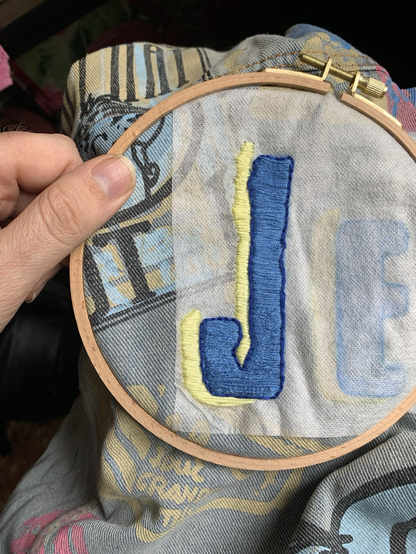 A letter J made with embroidery on a denim cloth. The letter is blue and it has a yellow shadow on the left. You can see the fingers of the perdón holding the clothe