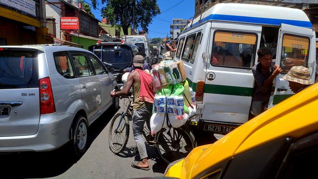 A cyclist, seen from behind, pushes his heavily loaded bike between gridlocked cars in Antananarivo, Madagascar. The bike's rear rack is stacked with items for sale in a neighborhood épicerie, including toilet paper, dishwashing liquid, and bags of rice.