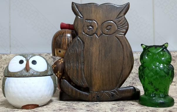 A photograph of three owl figures standing side-by-side on a kitchen counter. The left owl is a small, shourt, round condiment container from Japan with big eyes, gray sides, and a white breast. The center owl is made of three pieces for holding envelopes and other light papers. It is dark wood and has a somewhat crudely carved front. The third owl is short, emerald green, and made of bar glass. There appears to be a brown kokeshi doll peaking out from behind the center owl, but I deny it.