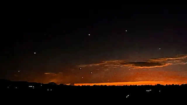 A dark evening sky with a bit of red sunset left near the horizon, and white dots hovering here and there: a photograph of drones over New Jersey.