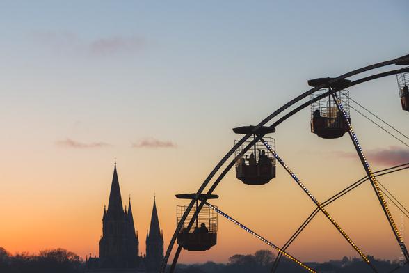 A silhouette of a Gothic cathedral with spires against an orange and blue sunset sky, with a Ferris wheel or observation wheel in the foreground. The wheel's gondolas are illuminated by LED lights, creating a striking contrast with the darkening sky.