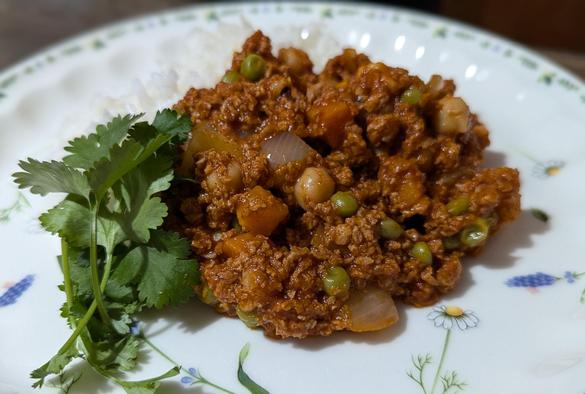 a plate of white rice and vegan keema curry, with some cilantro garnish on the side. the curry has textured vegetable protein, chickpea / garbanzo beans, black-eyed peas, green peas, carrots, onion, garlic, and ginger. it is all sitting on a white china plate that has flower graphics on it.