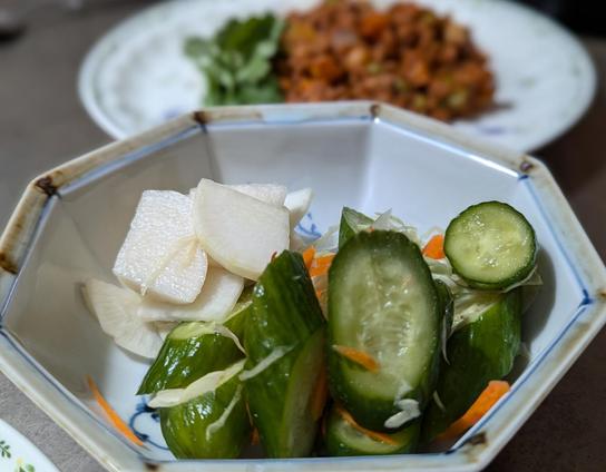 an octagon-shaped bowl of tsukemono (Japanese style pickles). Behind the bowl, out of focus, is a plate of vegan keema curry. In the foreground are large cut cucumber pickled with thinly sliced cabbage and carrot. Behind the cucumbers are cross-section slices of daikon, sweetly pickled.