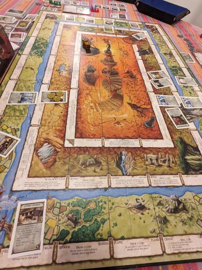 A colorful board game is set up with cards, tokens, and dice placed on the table. The board features a fantasy-themed design with various regions, paths, and spaces marked for gameplay.