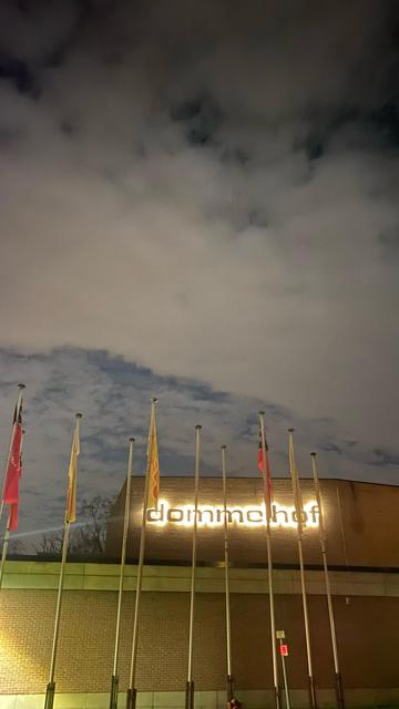 we see a brick building with flagpools in front of it. Another level of the building holds the name of the place 'Dommelhof', the letters have lights behind them so it can be read in the dark.
Above it all we the very cloudy evening sky.