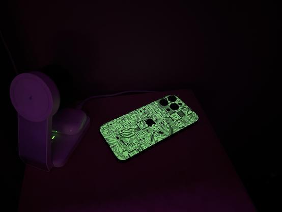 Telefon leżący plecami do góry z fluorescencyjną winylową naklejką. Obok ledwie widoczna ładowarka. Zdjęcie robione w ciemnych warunkach dla lepszego uchwycenia efektu fluorescencji.