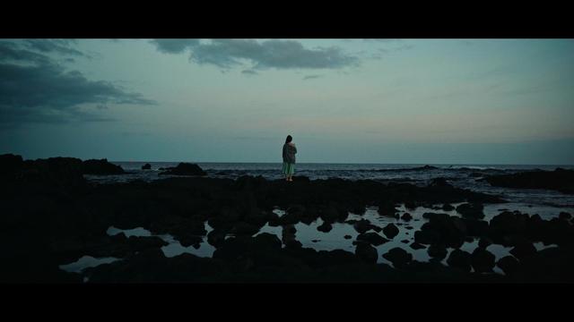 Eine Szene aus dem Kurzfilm "Santa Rita Dream". Eine Frau, mit dem Rücken zur Kamera, blickt in der Dämmerung auf das Meer hinaus.