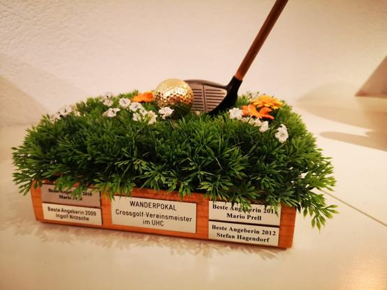 Unser Wanderpokal für den Vereinsmeister. Eine breite Holzplatte, auf der eine Kunstwiese mit Blümchen befestigt ist und rings rum am Rand, die Gravurschilder der Vereinsmeister. Oben drauf ist noch ein Stück eines Golfschlägers befestigt