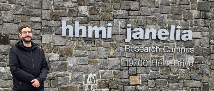 Photo of Peter Kraker in front of a stone wall with a sign saying: "hhmi | janelia Research Campus, 19700 Helix Drive"