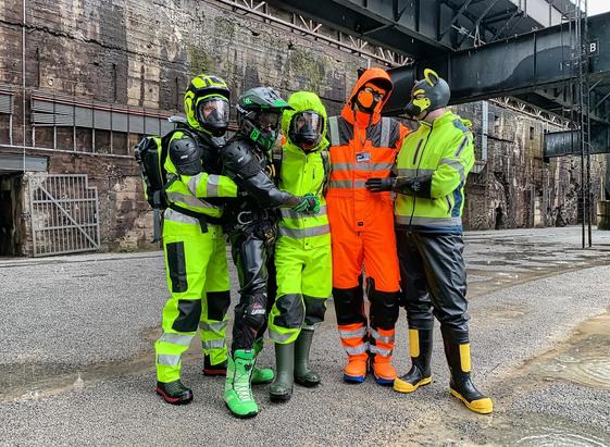 An old industrial complex with a large stone building with metal constructions in the background. The ground is sandy and wet, with several rain puddles and thick rain drops everywhere.
With some distance before the building stands a group of five gearheads arm-in-arm. On the far left Adensy in his yellow hi-vis raingear with MX chest protector, rubber boots, gas mask, helmet with ear protectors and a raingear backpack. He holds Hiscal from behind who stands next to him. Hiscal wears full-rubber with a belt harness, green snowboots, thick black-green gloves and a black-green MX helmet with goggles. To his right stands Exodiver in yellow hi-vis raingear with green rubber boots and a gas mask with the yellow raingear hood on top. Hiscal rests his right hand on the belly of Exodiver. Next stands the fox in his orange hi-vis one-piece boilersuit with his black-orange puppy mask and boiler suit hood on top, and orange rubber boots. To the far right stands Johnny the Buck in a yellow hi-vis jacket and black raingear pants and black-yellow rubber boots, black rubber gloves and his black-yellow puppy mask. He rests his left hand on the belly of the fox. All are facing the camera, except for Johnny the Buck who looks to the left at the group.
