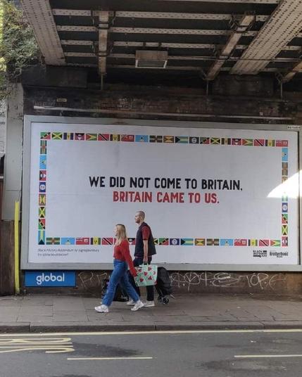 A billboard under Brondesbury Bridge in London, designed by Greg Bunbury for Black Outdoor Art and Black History Month 2022. The artwork features the phrase 'We did not come to Britain. Britain came to us,' surrounded by Caribbean flags, addressing the UK's colonial past and Windrush migration.