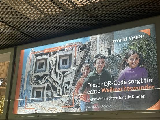 Photo of a campaign poster by »World Vision«. Smiling Children in front of a destroyed building, which through the use of generative AI is turned into a QR code. The slogan is: »Dieser QR-Code sorgt für echte Weihnachtswunder« (»This QR code creates real Christmas miracles«)