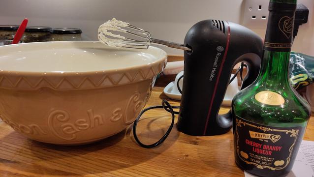 Mixing bowl with electric whisk stood on it's butt, creamy whisks hanging over the bowl. Next to it is a bottle of cherry liqueur.