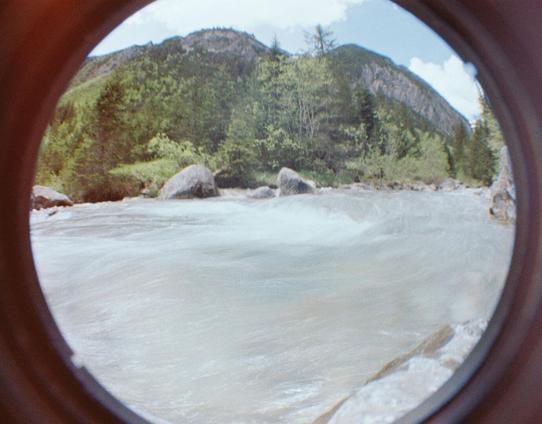 Ein ruhiger Flussabschnitt mit umliegendem Wald und Gebirge. Die hellen Töne des Himmels spiegeln sich leicht im Wasser, während die Landschaft durch die Vignettierung des Objektivs eingerahmt wird.
