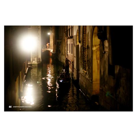 A nighttime scene in Venice, Italy, captures a gondolier guiding a gondola through a narrow, glowing canal. Light spills dramatically from a nearby building, reflecting across the water and casting surreal, rippling patterns. In the distance, figures stand on a small bridge, their silhouettes soft in the haze of a street lamp. Photo taken by Joseph Bautista.