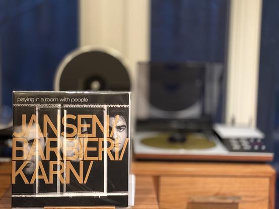 Jansen/Barbieri/Karn - Playing in a Room with People LP cover.

Three vertical strips with black and white photos of the three primaries, in that order. Title at the top, and the whole thing overlaid with semi-transparent gold letters with the participant names.

The gold LP plays on a vintage, restored Beogram 8002 linear-tracking turntable to the right.