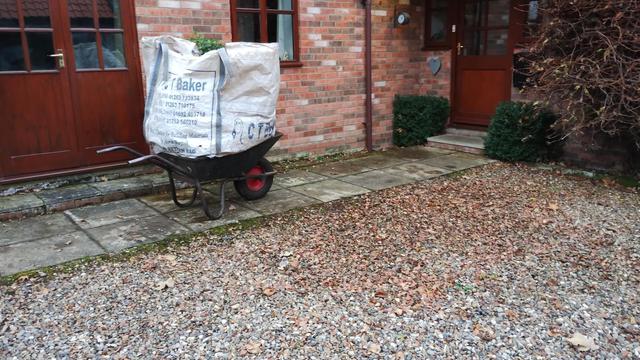 Paving slabs form a path to the front door of a house. There's a wheelbarrow on the path loaded with a bag full of leaves.