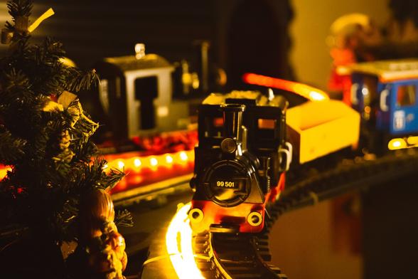 A festive toy train set illuminated with warm yellow lights runs along a miniature track. The black and red locomotive with the number "99 501" is in focus, pulling yellow and blue carriages behind it. To the left, part of a decorated Christmas tree with ribbons is visible, adding to the holiday ambiance.