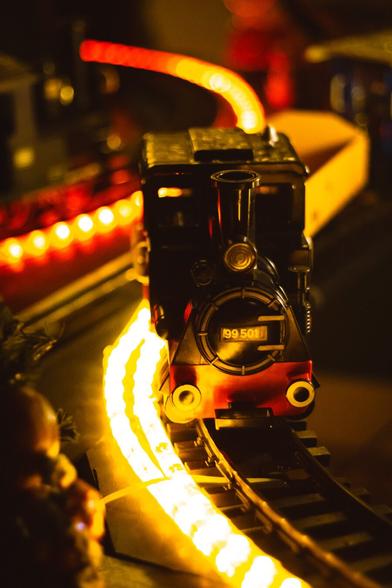 A close-up view of a toy train on a miniature track, featuring a black and red locomotive with the number "99 501" prominently displayed. The train is surrounded by glowing yellow lights, creating a warm and festive atmosphere. The blurred background adds depth, with hints of holiday decorations and the train’s carriages.