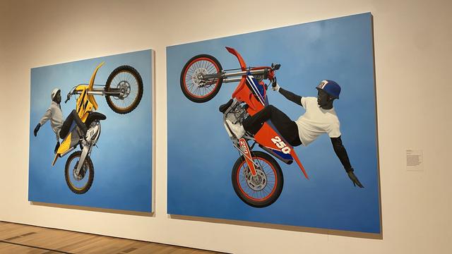 The two motorbike paintings by Amy Sherald taken from a slight angle standing right of the two pieces.  The piece on the left is a yellow dirtbike popping wheelie and the piece on the left is a red dirtbike popping a wheelie.  The two bikes are facing each other with a symmetrical arrangement. 
Description of Amy's work is in the alt text of the 3rd pic attached.
