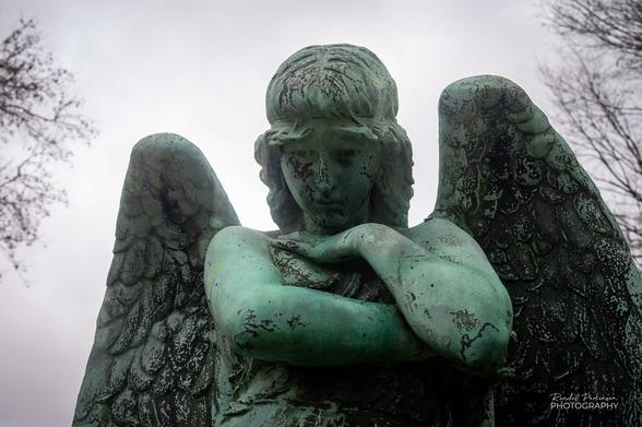 Photo showing the upper body of a bronze statue of an angel looking down at the family graves in front of the memorial.
