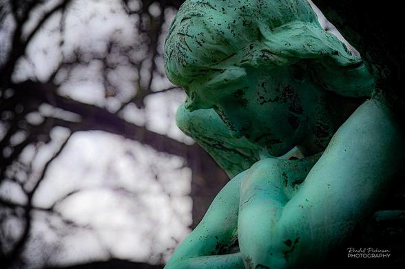 Side few of the head and upper body of a bronze statue of angel looking down at the ground in front of a large family cemetery monument.