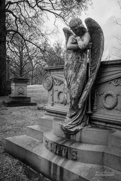 Black and white image of a bronze angel standing at the top step of a stone cemetery monument marked with the family name of Hilts.