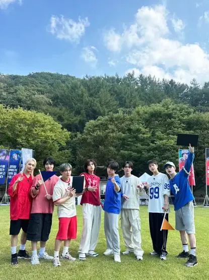 Promo still from skz code field day episode. Red team: Felix, I.N, Changbin, Hyunjin. Blue team: Han, Lee Know, Seungmin, Bang Chan. Their team banners are visible in the background. Strangely, the red team has blue banners, and the blue team has red banners.