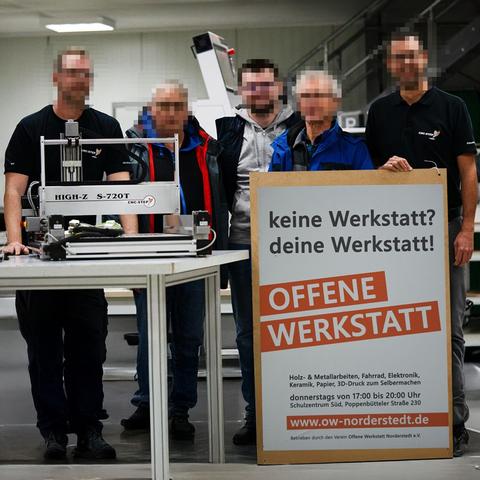Abholung unserer neuen Fräse bei CNC-Step in Geldern. Auf einem Tisch steht die CNC-Fräse, daneben ein Werbeplakat der Offenen Werkstatt Norderstedt mit dem Slogan: "keine Werkstatt? deine Werkstatt!". Dahinter stehen 5 Männer.