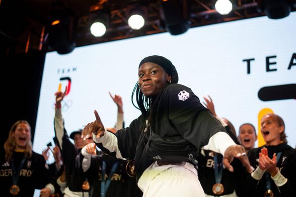Nicole Anyomi im Bildvordergrund, sie hat die Haare am Oberkopf zusammen und weiter unten in Zöpfen offen, trägt ein schwarzes Oberteil mit weißem Bundesadler am Ärmel und eine weiße Hose, sie tanzt. Hinter ihr in der Unschärfe Spielerinnen der Nationalelf, vor einer weißen Leinwand, auf der das "Team Deutschland"-Logo der Olympischen Sommerspiele in Paris zu sehen ist. Credit: IMAGO/Eibner