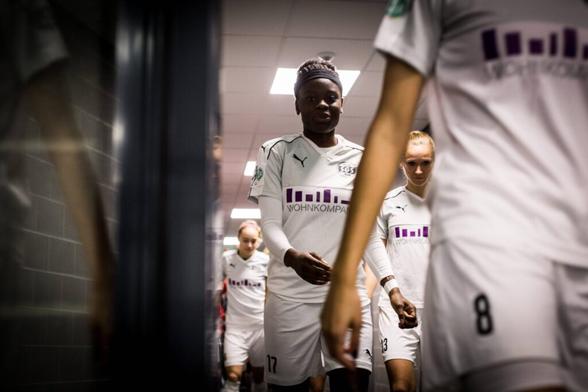 Eine junge Nicole Anyomi im weißen Trikot der SGS Essen läuft mit ihren Mitspielerinnen einen Gang entlang. Am rechten Bildrand eine offene Tür, durch die die Spielerinnen gerade gehen, vorne im Bild der Körper der vordersten Spielerin in der Unschärfe, dahinter Anyomi. Credit: IMAGO/Nordphoto