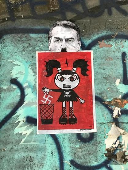 Streetartwall. Two self-painted paste-ups with a clear message have been stuck on a gray wall of graffiti. The bottom red image shows a fierce-looking black and white cartoon character, a little girl in a short black dress with a skull and crossbones print. She has two pigtails with skull hair clips and boots with red laces on. She is angry and throws a white swastika symbol into a garbage can. Above it is the second picture. It shows a half-drawn male head in black and white. It is the Brazilian ex-president Jair Bolsonaro. However, he has a black moustache like Adolf Hitler. 
Note: Radical Playground has produced these half heads with Hitler moustaches for a number of leading politicians.