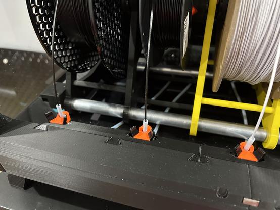 This shows the interior of the dry box.  There are three filament spools.  (Actually there are five, but the other two are outside the photo.)  Each spool is feeding filament into a PTFE tube.  The tubes are held by orange printed inlets, and the inlets are locked into the trapezoidal slots of the adapter pieces.  The adapter pieces have five slots, and on the other side they have six trapezoidal tabs that fit into the six slots in the dry box.