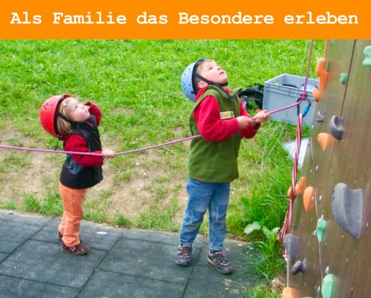 Zwei kleine Kinder mit Helm am Boden einer Kletterwand, die ein Sicherungsseil halten
