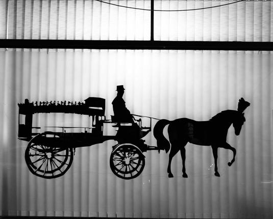 A man sitting in a horse-drawn carriage, seen in the window of a funeral parlor.

Ein Mann sitzt in einer Pferdekutsche, gesehen im Fenster eines Bestattungsinstituts.