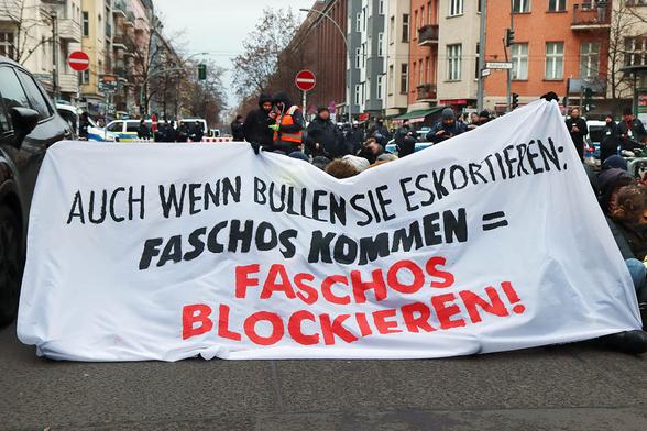 Das Foto zeigt eine Sitzblockade, verdeckt mit einem Transparent mit dem Text: "Auch wenn Bullen sie exkortieren: Faschos kommen = Faschos blockieren!"