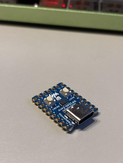 An electronic circuit board with a blue surface, featuring a USB-C connector, two buttons, and a small LED. The board is positioned on a light-colored surface with a blurred background.