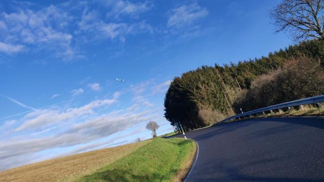 Blauer, leicht bewolkter Himmel, kleines Bergsträßchen und ein Jet am Himmel