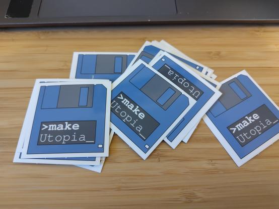 Es liegen mehrere Sticker auf einem Holztisch. Die Sticker sind quadratisch mit einer Seitenlänge von 5cm und haben eine blaue Diskette abgebildet auf der ">make Utopia_" geschrieben steht.