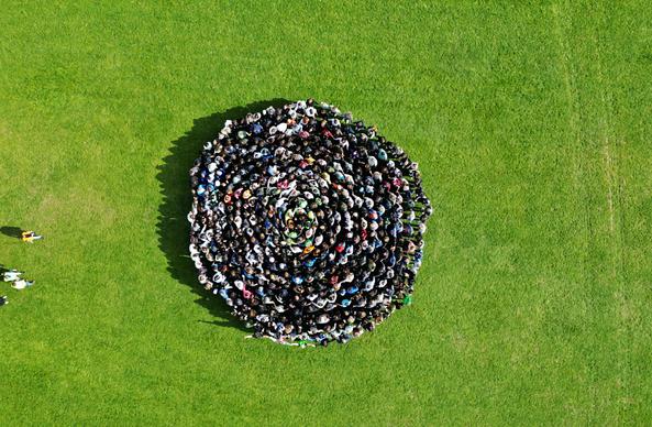 470 junge Menschen sich an den Händen zusammenfassend eng zusammen gerollt wie eine riesige Zimtschnecke. Auf einer grünen Wiese mit einer Drohne aus der Luft fotografiert.