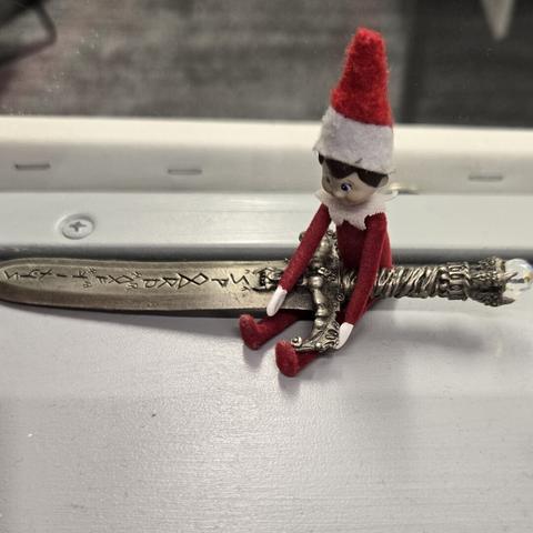 Photograph of a tiny Elf on the Shelf sitting on a ledge with a large (comparatively) sword shaped letter opener with runes all across the blade.