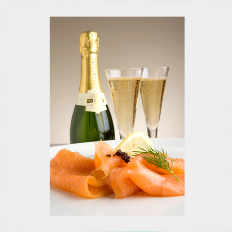 In the foreground, slices of smoked salmon garnished with dill, lemon, and caviar. In the background a bottle of champagne and two glasses of champagne.