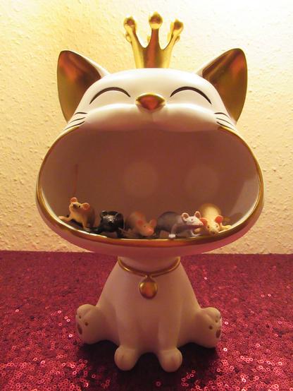 Deko-Katze, deren weit geöffnetes Maul als Ablage dienen kann. Darin fünf kleine Spielzeugmäuse. Sie ist weiß, mit goldenen Details und goldenem Krönchen.