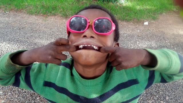 A Malagasy boy making a fish face, wearing oversized sunglasses with pink frames. The sunglasses are humorously repaired at the bridge with a rubber band