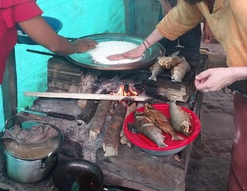 two people are pressing cassava starch (tapioca) onto a large , flat frying pan over a wood fire. There is freshly grilled fish in the foreground and a turquoise tarp wall in the background.