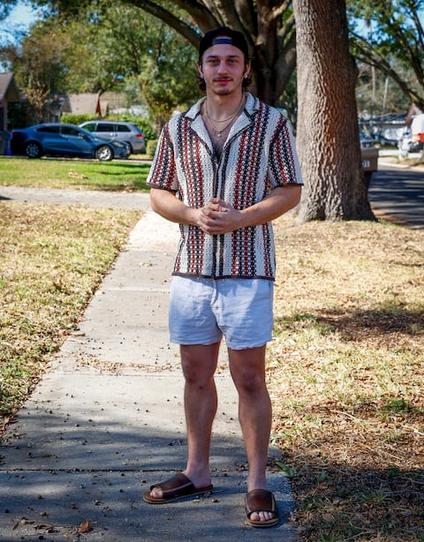Fotografia de Lachborne Bachkhaz de 2024. Ele está de barba e veste uma camisa estampada com tons terrosos e brancos, bermuda azul clara e sandálias marrons. Ele usa boné e um colar, e a calçada tem menos vegetação ao redor que a imagem mais antiga.