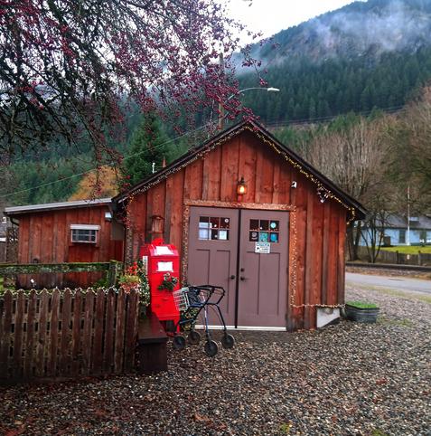A charming rustic wooden building set against a backdrop of misty forested mountains. The building has a warm, weathered appearance with vertical wood paneling and brown double doors adorned with small square windows. Festive string lights outline the roof, creating a cozy atmosphere. A bright red mailbox and a shopping cart walker rest near a wooden picket fence to the left of the building, adding character. The foreground features a gravel path, and the scene is framed by branches of a tree with red berries. The setting conveys a tranquil, small-town vibe.