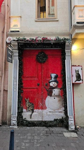 Streetart. A beautiful mural with a Christmas motif was painted on the old door of a modern clothing store. The red-painted door forms the background for a large, smiling snowman with a top hat and tartan scarf. A beige cat sits next to him and looks at him with interest. Another cat is painted on the letterbox next to the door. The door itself is decorated with a Christmas tree arch and a Christmas door wreath, above which there is lighting with fir greenery. 
Info: The door was painted last year for Advent and looked different the rest of the year. Only the cat remained and the rest of the door was painted half pink and half white. This year the snowman is back, a deer head and many other things have been added, but unfortunately the cat has disappeared. What a pity.