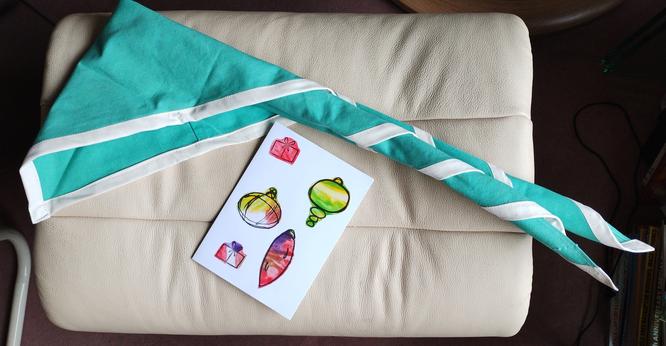 A pale turquoise green scout neckerchief with white edging rolled up ready to wear, laid on a footstool.
Beside it is a Christmas card featuring cut-out baubles and presents coloured by crayons.